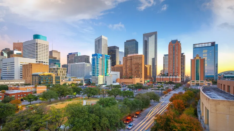 City skyline at sunset with high-rise buildings, green trees, and light trails on streets.