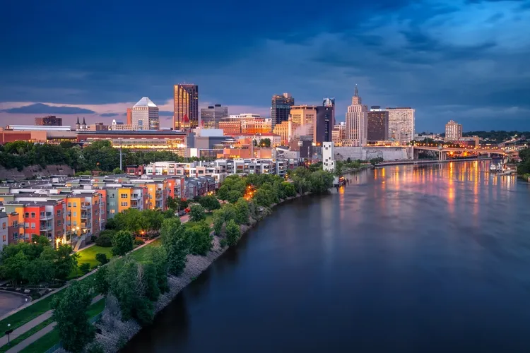 Aerial view of a city skyline at dusk with illuminated buildings and a riverfront.