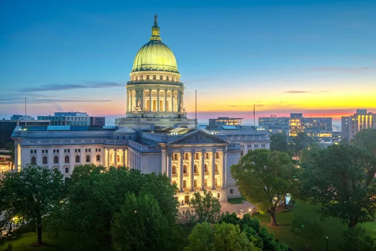 Illuminated classical government building with large dome at sunset, surrounded by trees and cityscape.
